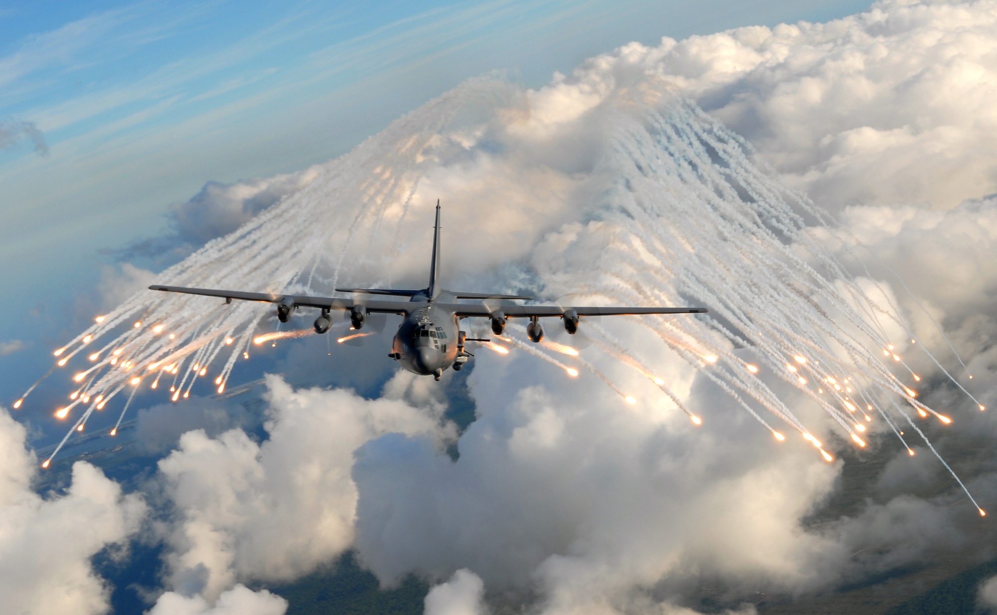 AC-130 aircraft releasing a broad fan of countermeasure flares in a blue-gray daylight sky